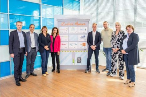 Groepsfoto van de acht bestuurders die het commitment voor Zwitch Almere ondertekenden, poseerend rondom een paneel met logo's . In het midden, v.l.n.r.: P. Rotering (Zorggroep Almere), M. Seelen (Flevoziekenhuis), N. Nieuwenhuizen (gemeente Almere), N. Tellegen (Flever), S. van de Sant (Woonzorg Flevoland), Y. Zwijnenburg (Stichting Moving Up), J. Veltman (directeur ROC van Flevoland) en L. Voorhorst (Triade Vitree) .