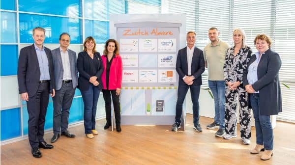 Groepsfoto van de acht bestuurders die het commitment voor Zwitch Almere ondertekenden, poseerend rondom een paneel met logo's . In het midden, v.l.n.r.: P. Rotering (Zorggroep Almere), M. Seelen (Flevoziekenhuis), N. Nieuwenhuizen (gemeente Almere), N. Tellegen (Flever), S. van de Sant (Woonzorg Flevoland), Y. Zwijnenburg (Stichting Moving Up), J. Veltman (directeur ROC van Flevoland) en L. Voorhorst (Triade Vitree) .