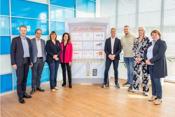 Groepsfoto van de acht bestuurders die het commitment voor Zwitch Almere ondertekenden, poseerend rondom een paneel met logo's . In het midden, v.l.n.r.: P. Rotering (Zorggroep Almere), M. Seelen (Flevoziekenhuis), N. Nieuwenhuizen (gemeente Almere), N. Tellegen (Flever), S. van de Sant (Woonzorg Flevoland), Y. Zwijnenburg (Stichting Moving Up), J. Veltman (directeur ROC van Flevoland) en L. Voorhorst (Triade Vitree) .